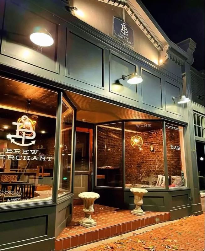 The Brew Merchant storefront at night, its anchor logo on the window and lamps lit above the entrance.