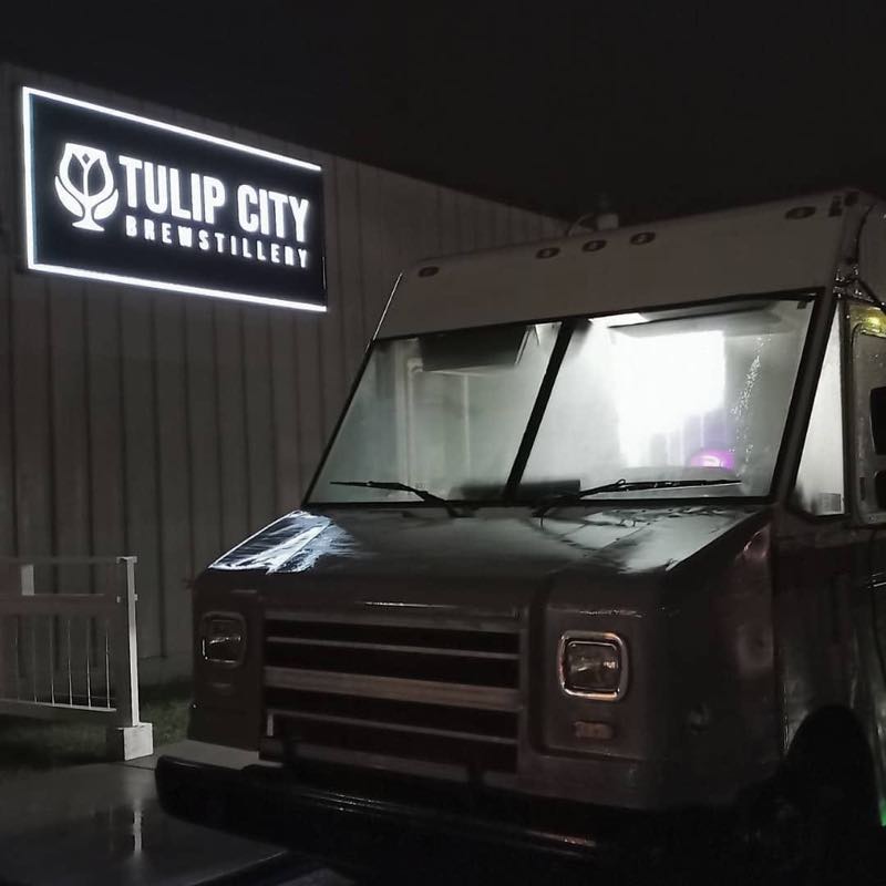 The lit Tulip City Brewstillery sign on the building wall at night, beside a parked food truck.